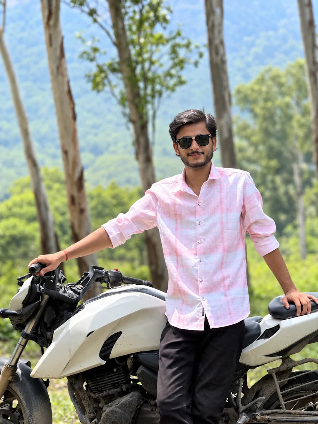 Suraj Mishra beside a white motorcycle in a wooded mountain setting